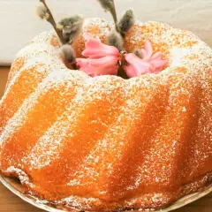 Puszysta babka jogurtowa oprószona cukrem pudrem, ozdobiona baziami i różowymi kwiatami. Idealna na wielkanocny stół.