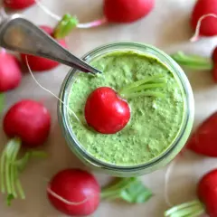 Słoik z zielonym pesto, ozdobiony rzodkiewką i otoczony innymi rzodkiewkami.