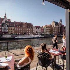 Gdańsk restauracje z widokiem na Motławę. Ludzie siedzą przy stolikach na drewnianym tarasie, podziwiając zabytkową zabudowę i pływające statki.
