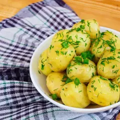 Młode ziemniaki w białej misce, posypane świeżym koperkiem i szczypiorkiem, na tle kratkowanej serwetki i drewnianego stołu.