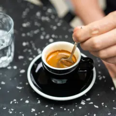 Ręka miesza łyżeczką aromatyczną kawę z ekspresu w czarnej filiżance. Obok stoi szklanka wody.
