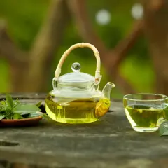 Szklany dzbanuszek i filiżanka z zieloną herbatą, obok świeże liście. Różne rodzaje zielonej herbaty zachwycają smakiem.