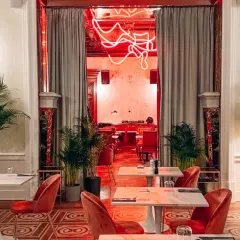 Eleganckie wnętrze z czerwonymi aksamitnymi krzesłami i marmurowymi stolikami. Neonowy napis w głębi sugeruje, że to najdroższa restauracja w Warszawie.