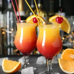 Dwa kolorowe, warstwowe drinki z pomarańczą i wiśnią, idealne na domowe drinki. W tle bar z butelkami i światełka.
