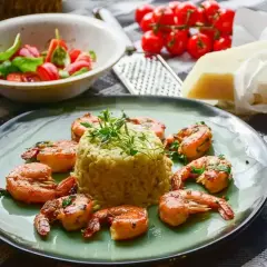 Krewetki z mlekiem kokosowym i curry na risotto, udekorowane ziołami. Obok miseczka z pomidorkami i bazylią, ser i tarka.