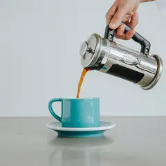 Ręka przelewa świeżo zaparzoną kawę z french pressa do turkusowej filiżanki.