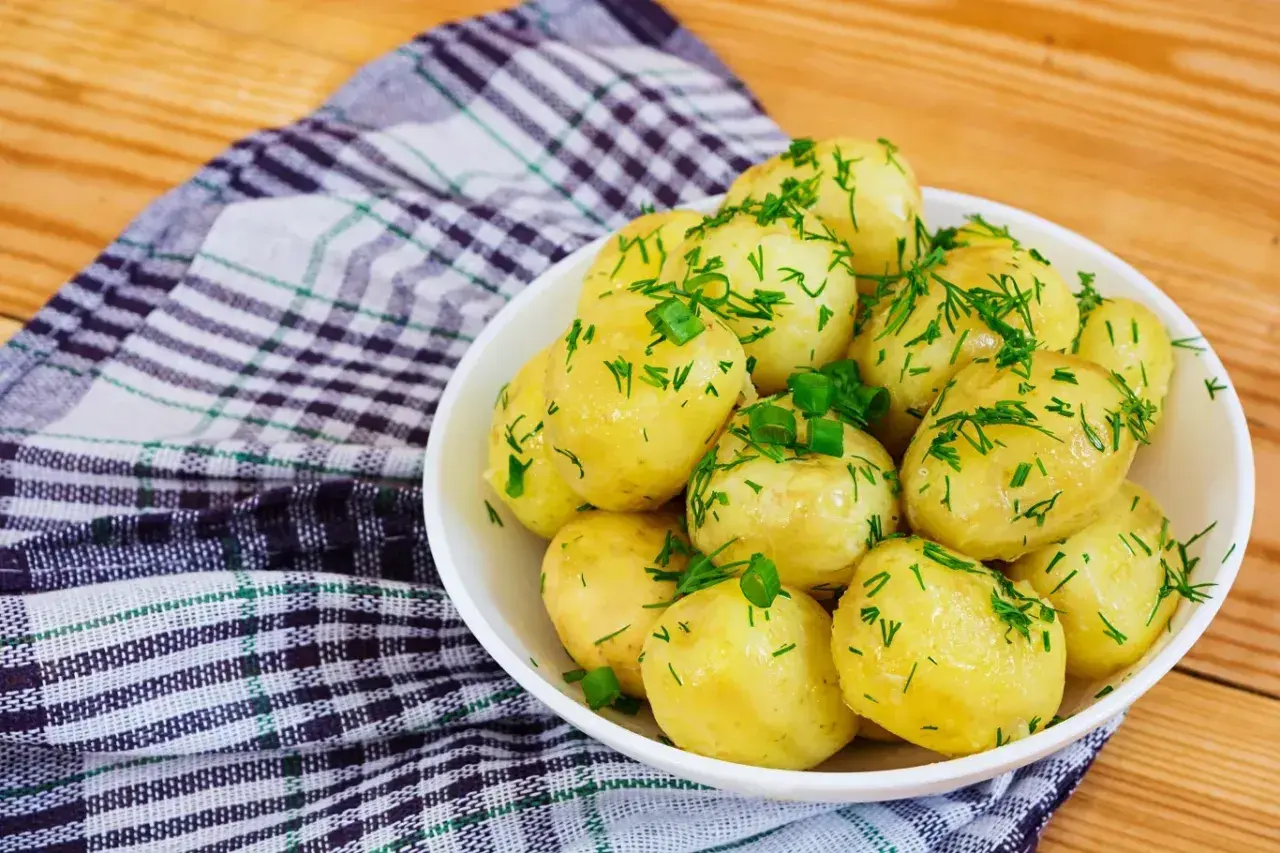 Młode ziemniaki w białej misce, posypane świeżym koperkiem i szczypiorkiem, na tle kratkowanej serwetki i drewnianego stołu.