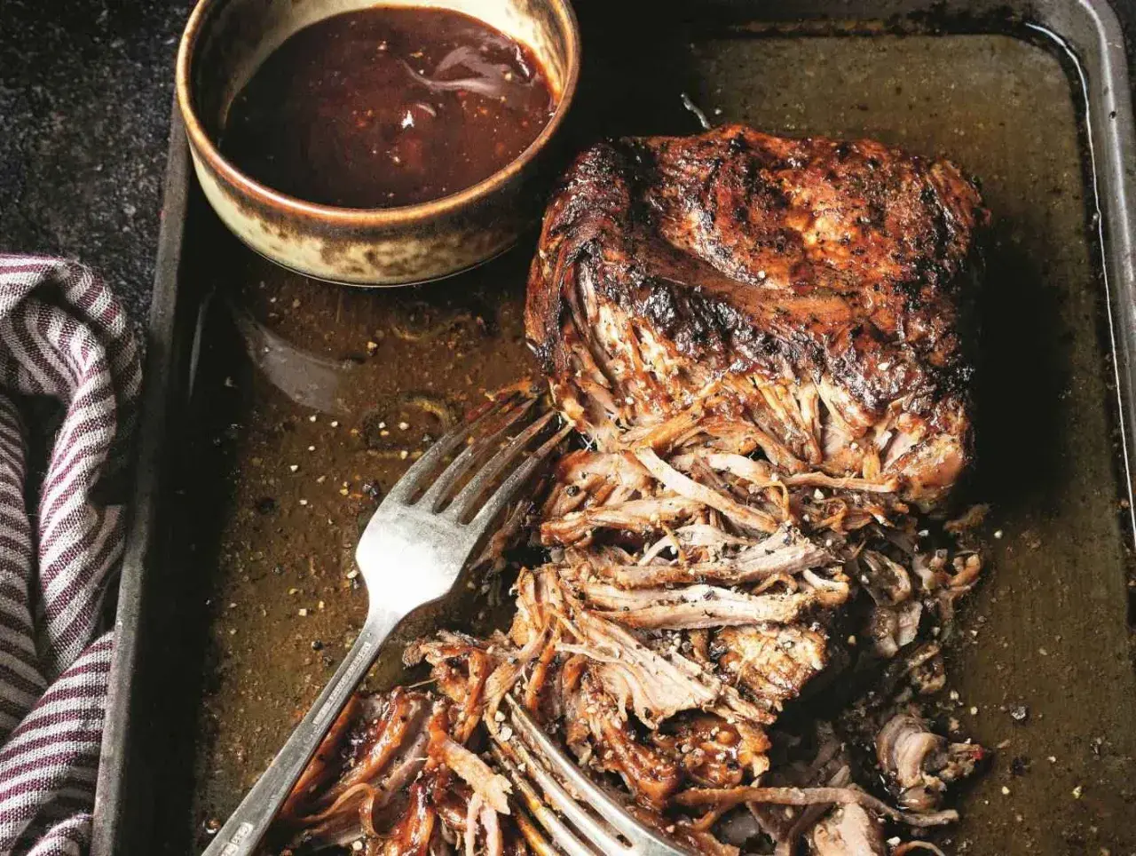 Soczysta, szarpana wieprzowina z sosem barbecue na blasze. Widoczne widelce ułatwiające rozrywanie mięsa.
