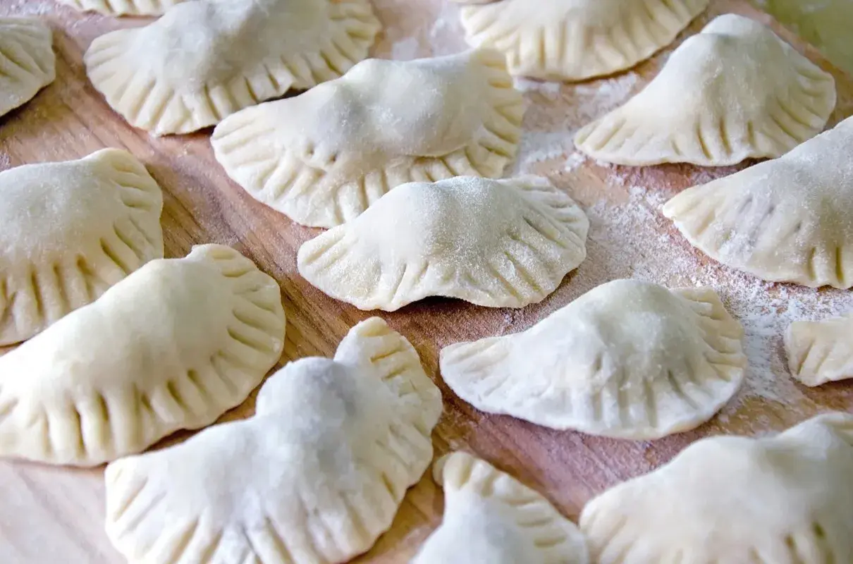 Surowe pierogi z kaszą gryczaną ułożone na drewnianej desce, posypane mąką.