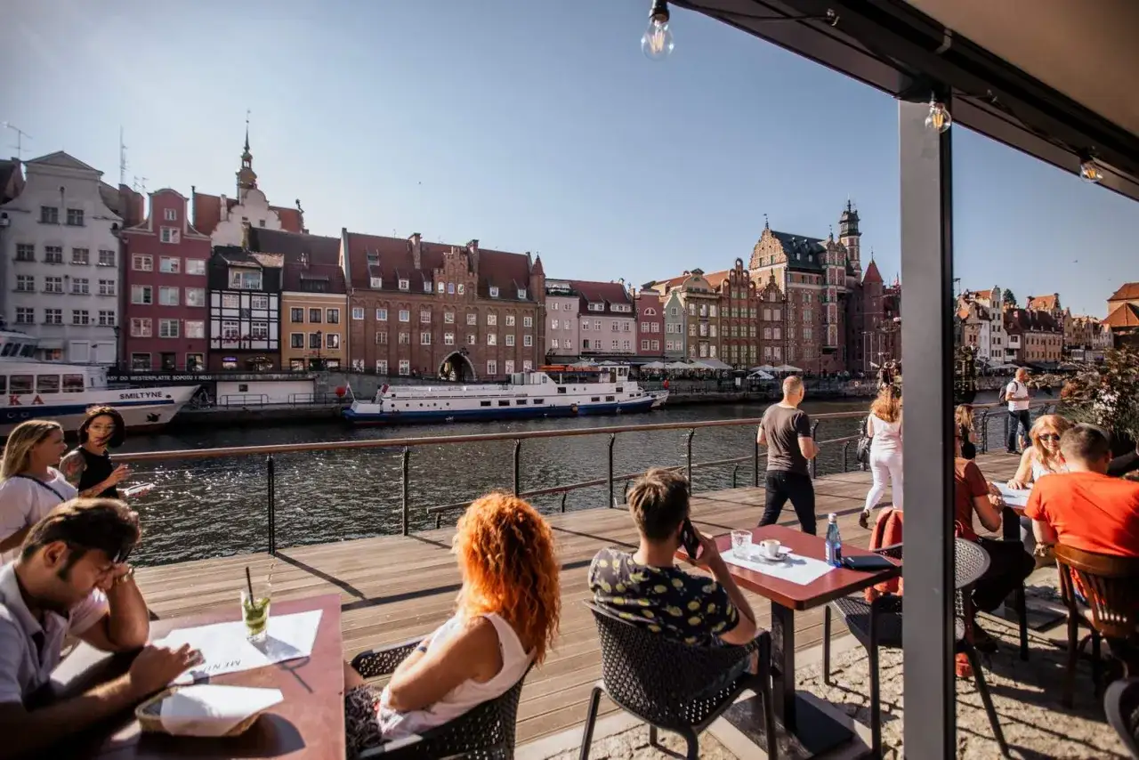 Gdańsk restauracje z widokiem na Motławę. Ludzie siedzą przy stolikach, podziwiając zabytkową zabudowę i pływające statki.