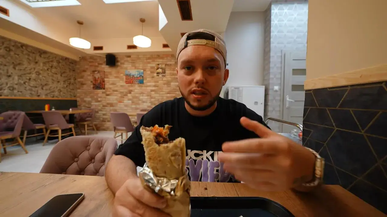 Mężczyzna z kebabem w ręku, prawdopodobnie najlepszy kebab warszawa. Wnętrze restauracji z ceglanymi ścianami i wygodnymi fotelami.