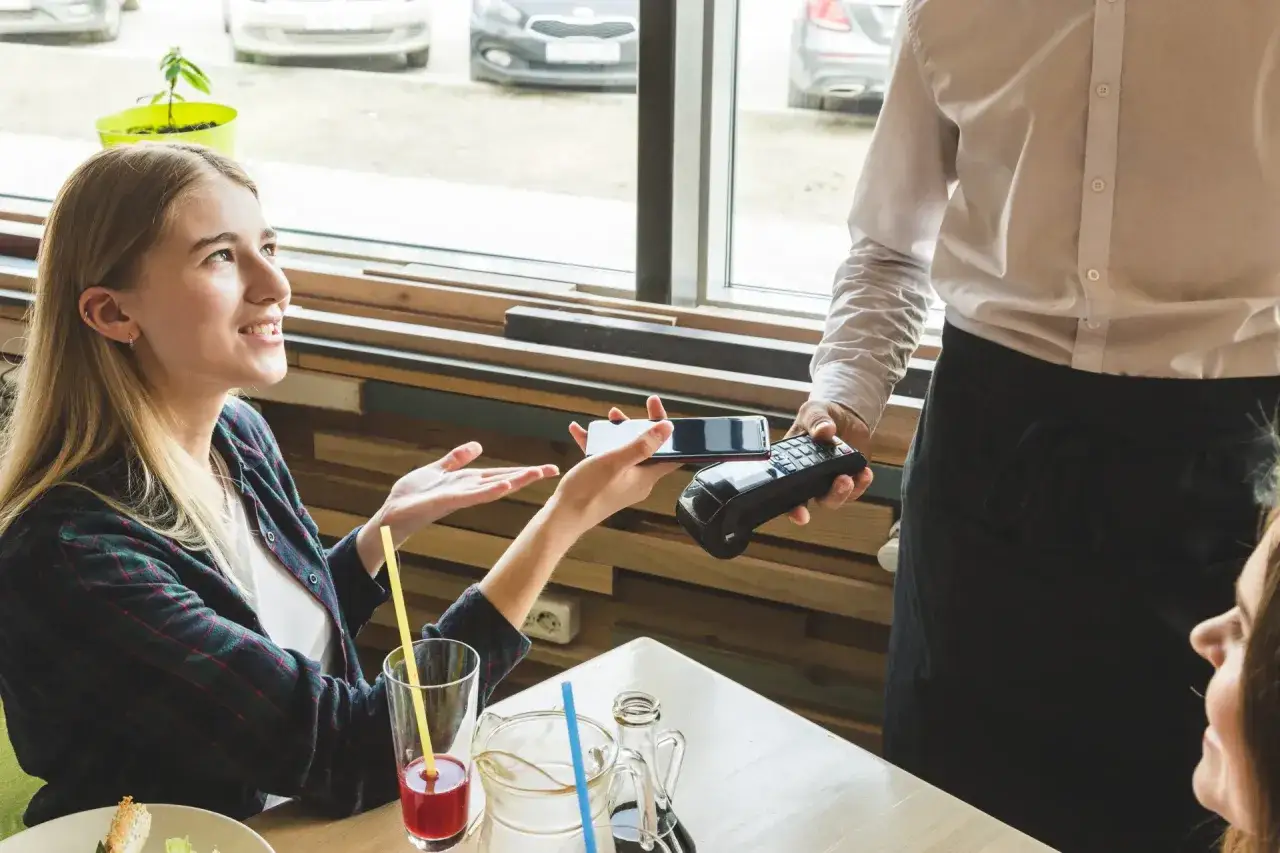 Uśmiechnięta dziewczyna płaci telefonem za posiłek, kelner trzyma terminal. Dzielenie rachunku w restauracji nigdy nie było tak proste.