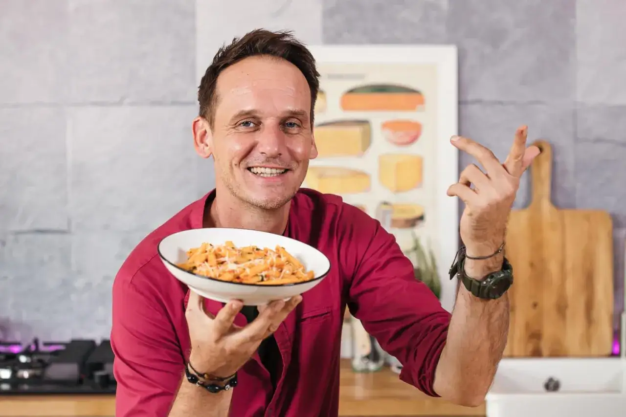 Pascal Brodnicki prezentuje talerz makaronu penne z sosem pomidorowym i parmezanem, uśmiechając się do widza.