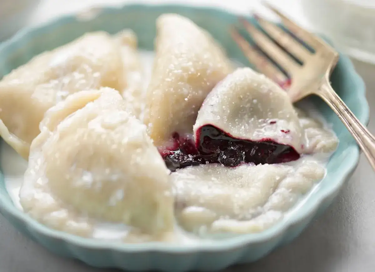 Pyszne pierogi z jagodami, polane śmietaną i posypane cukrem, czekają na widelcu.